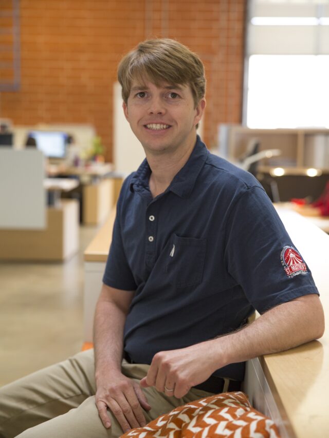 Headshot of man sitting on bench in an office wearing a blue polo shirt.