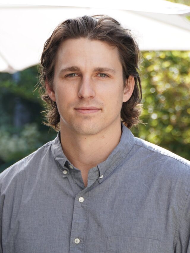 Headshot of man wearing gray shirt with greenery and umbrella shad in background.
