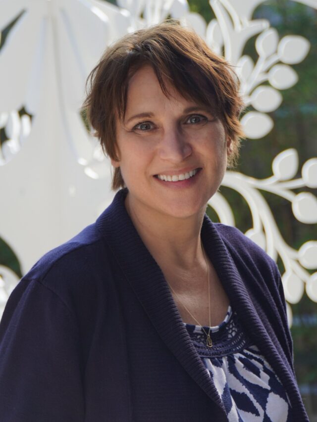 Headshot of woman wearing navy blue jacket and patterned top with a white sculpture and greenery in the background.