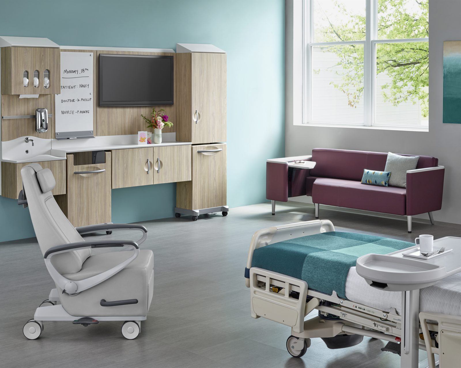 A vibrant exam room with a bed, recliner and sofa. Along the wall is casework with a sink run and storage. An open window provides warm light.