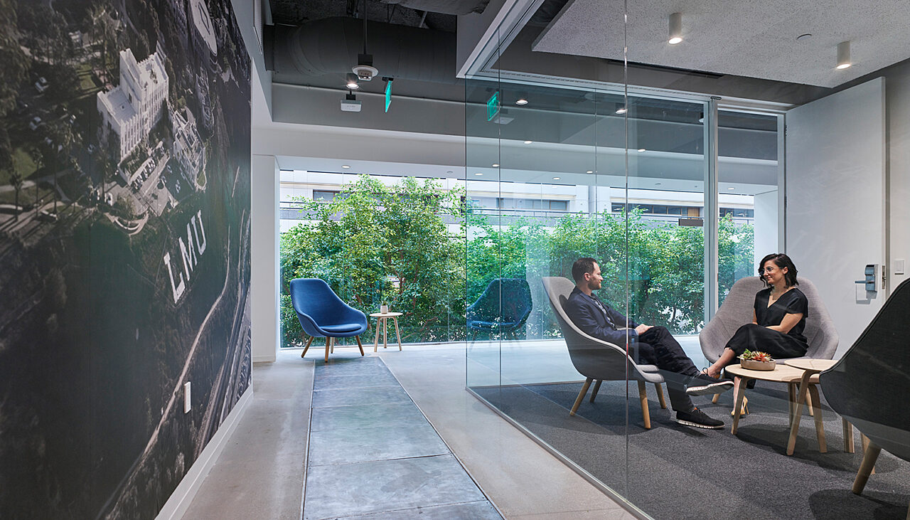 A small meeting room separated by a glass wall features a grey carpet, with a man and woman seated on grey lounge chairs next to a white coffee table.