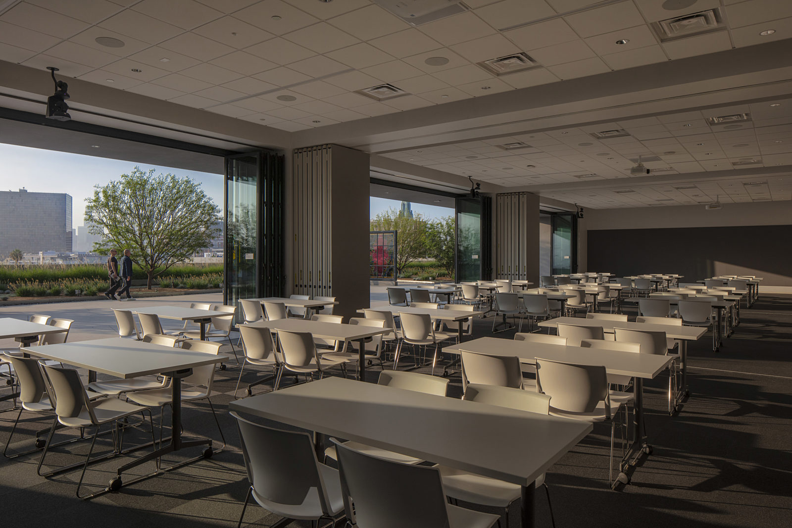 A spacious meeting room with stackable seating and tables, located beside an open walkway with outdoor views, perfect for modern interior design.