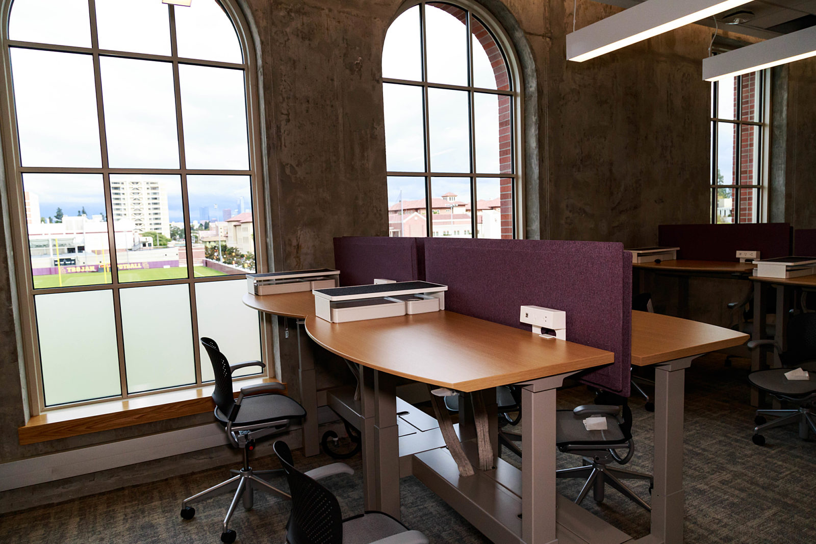 A huge workplace with several wooden desks and executive chairs. French windows with a magnificent view flank the desks.
