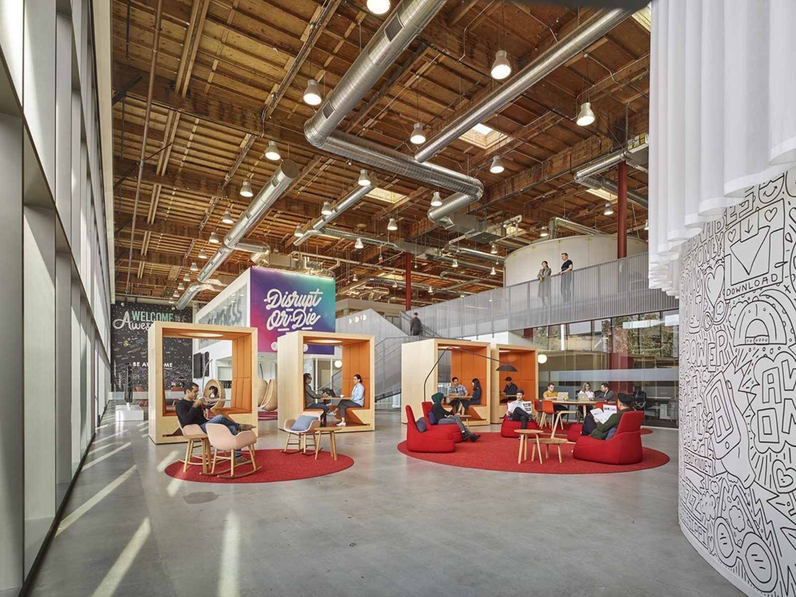An open space with high ceilings. Round rugs on the floor. Booths for small meetings or independent work. An interior bridge for access to second floor.