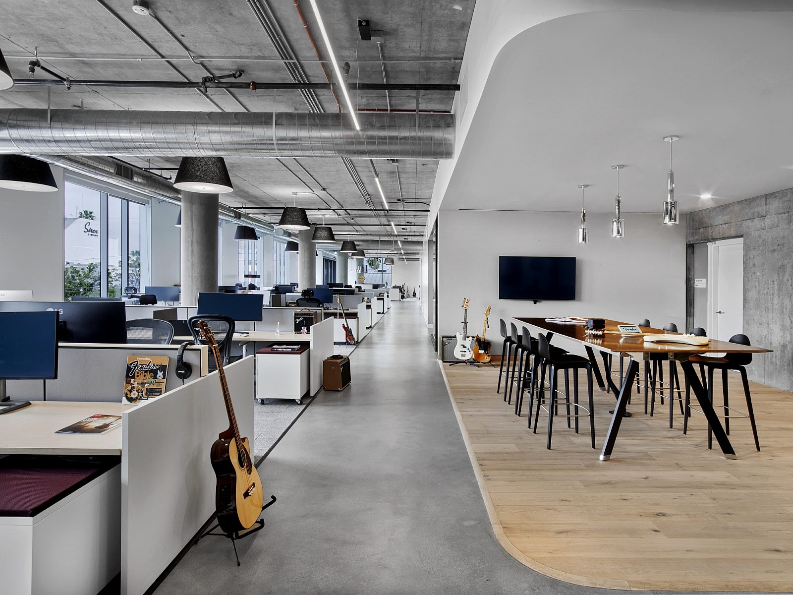 A muted workspace with cool colors. Guitars scattered throughout. The workstations line the left and on the right is a long high table with stools.