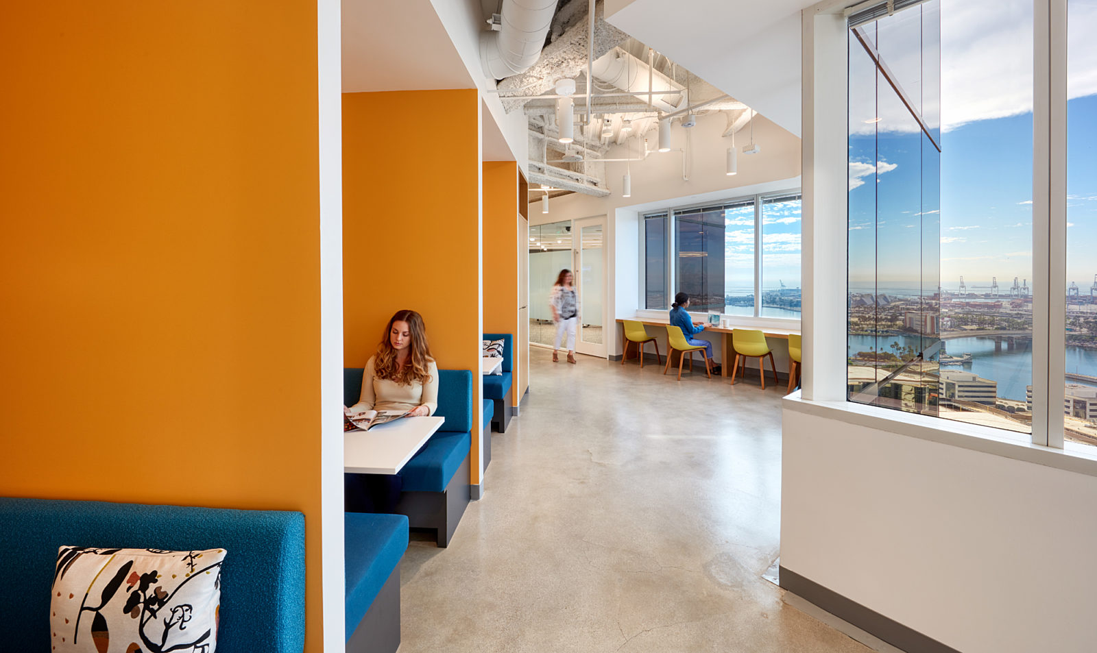 A mixed workspace featuring booths with colorful walls and upholstery and tables that line the wall on the right side overlooking a view of the city.