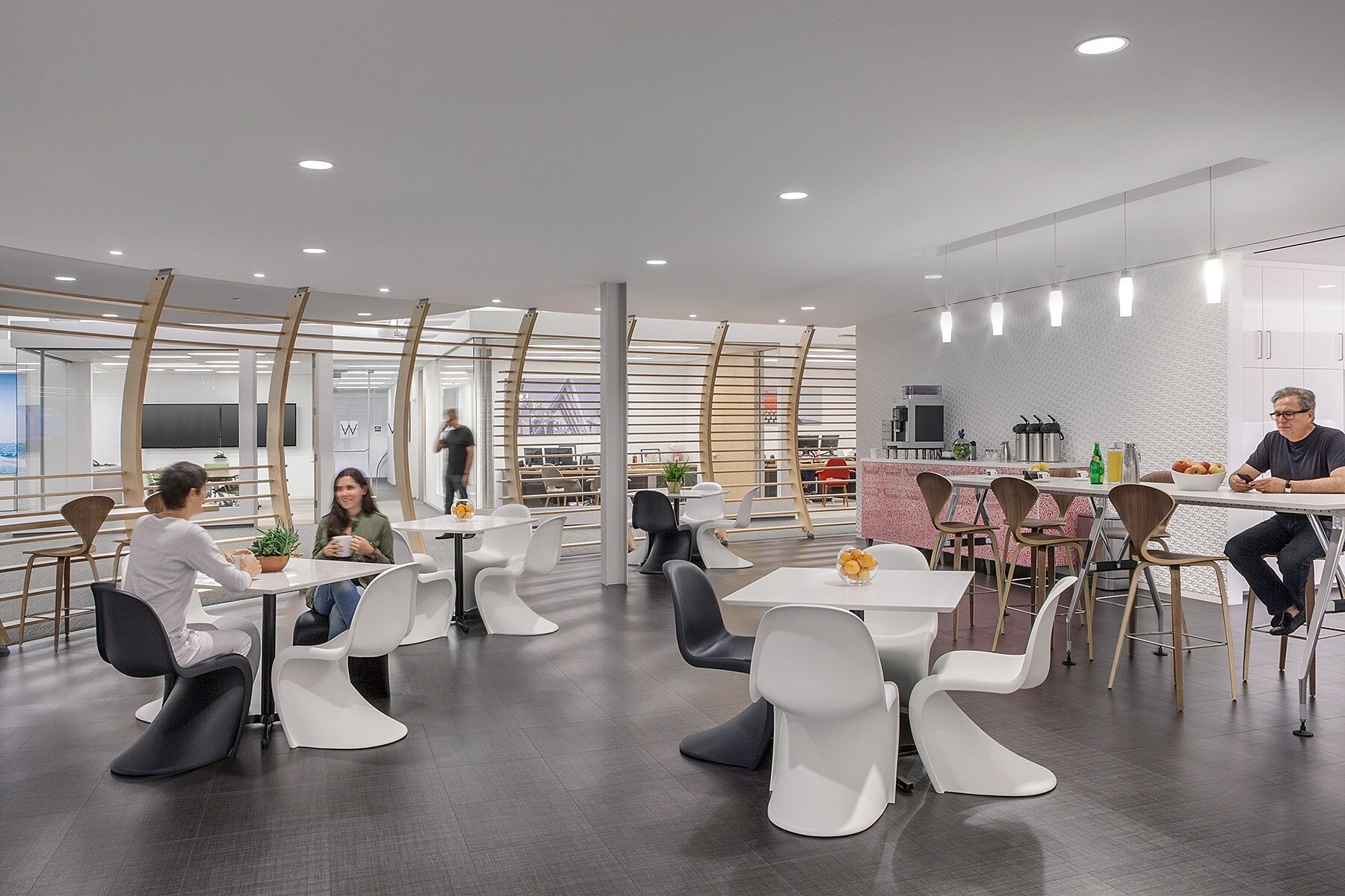 A modern cafeteria in a work setting. Cool colors featured on tables and chairs. High tables and stools line the background. The space features 4 people.