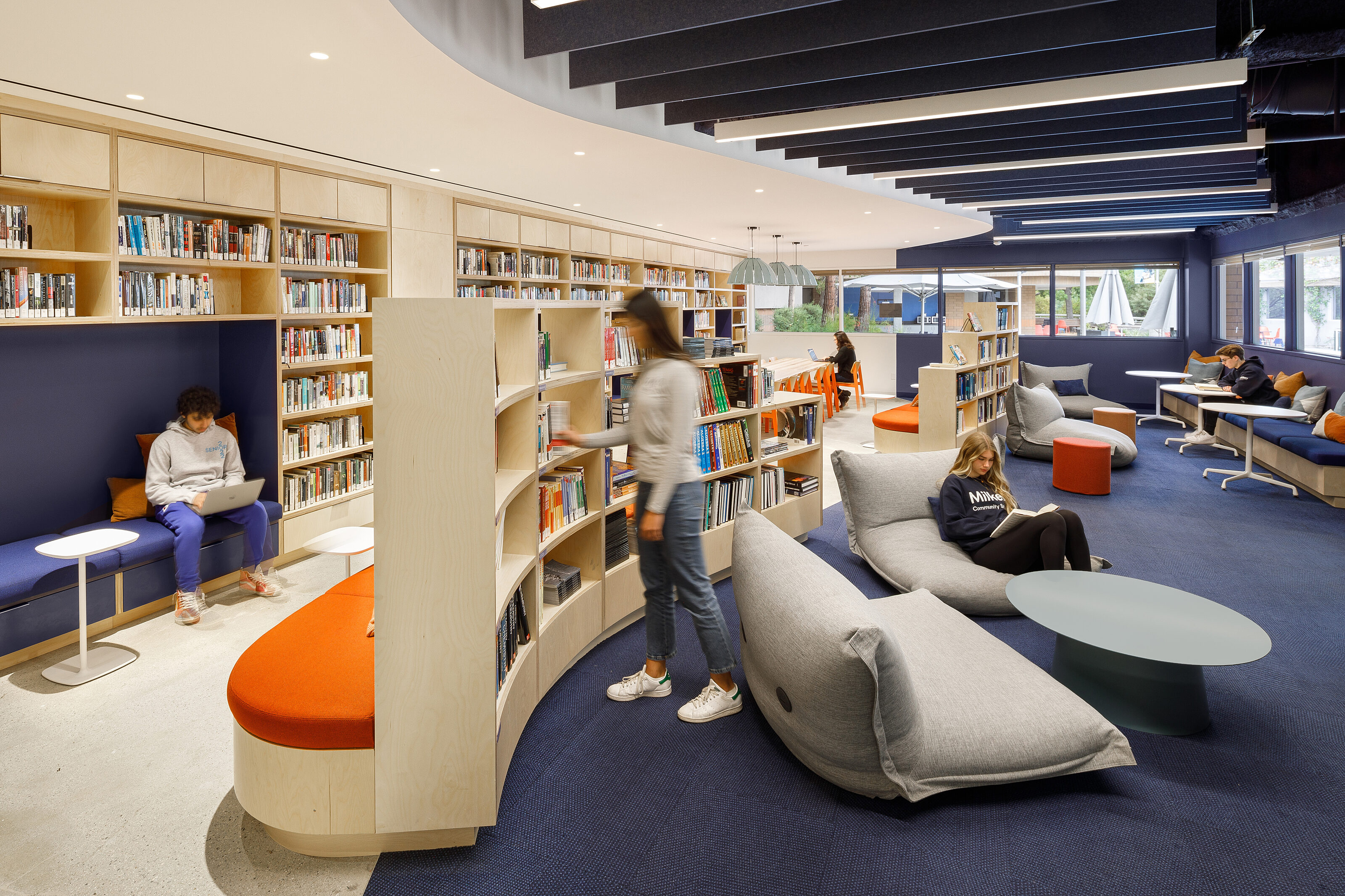 In the open reading area, custom bookshelf booths offer cozy seating along with lounge chairs and coffee tables, all situated on a dark blue carpet.