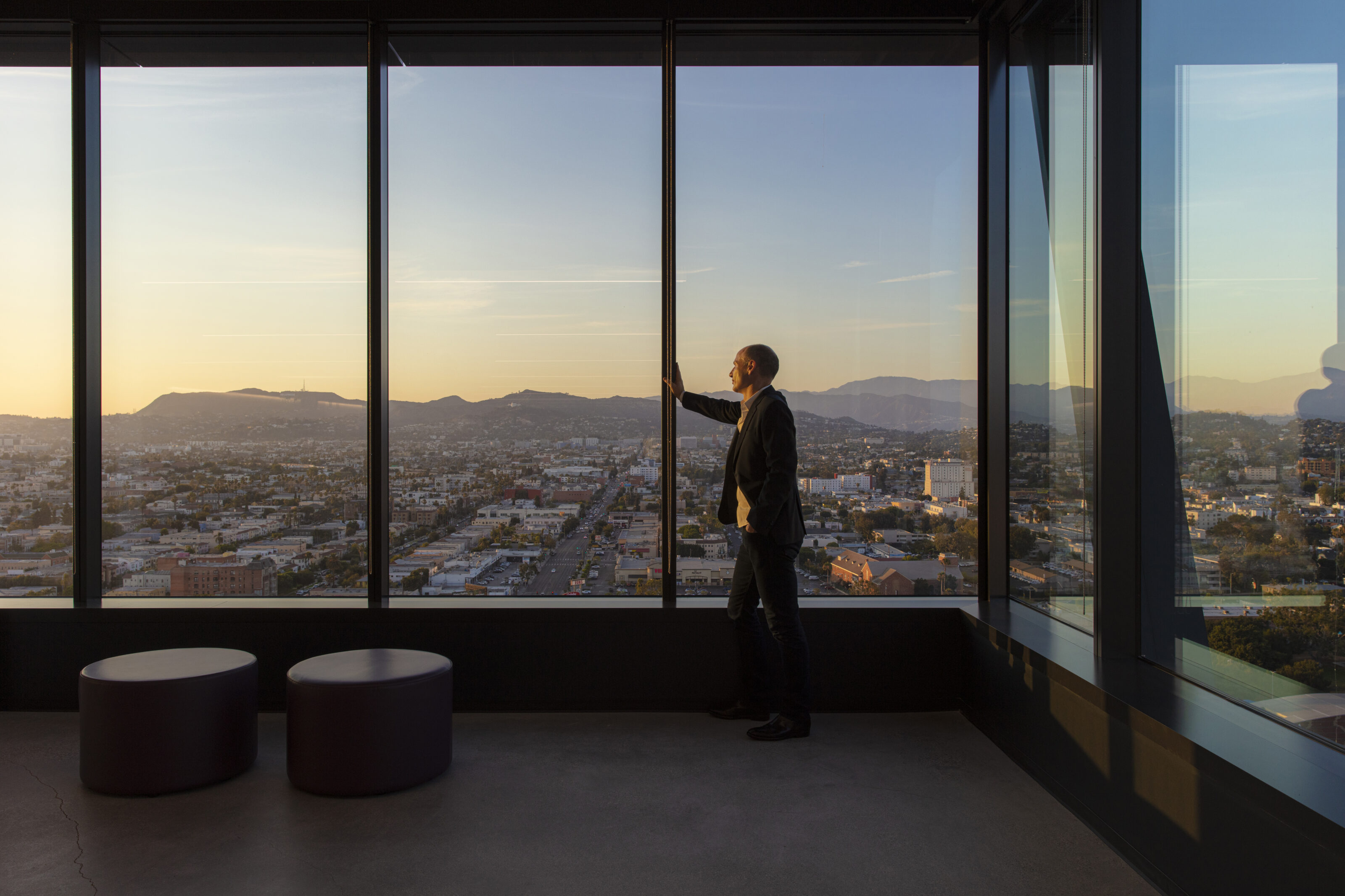 An area with French windows and stunning mountain views. A man is on his phone next to the ottomans that are positioned next to the windows.