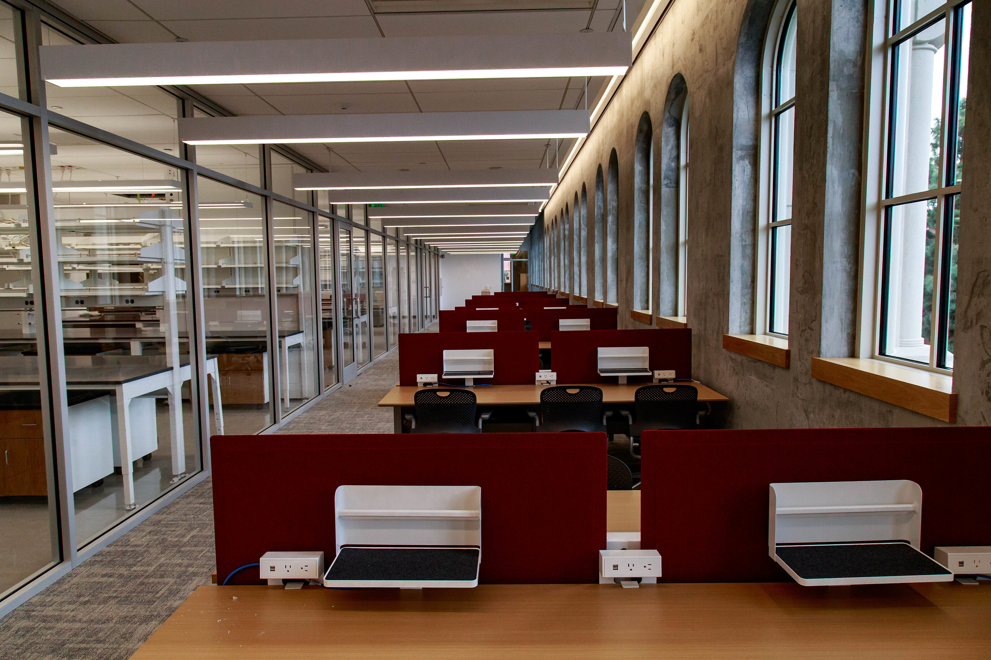A spacious open office with paired wooden desks positioned between French windows and a lab room.