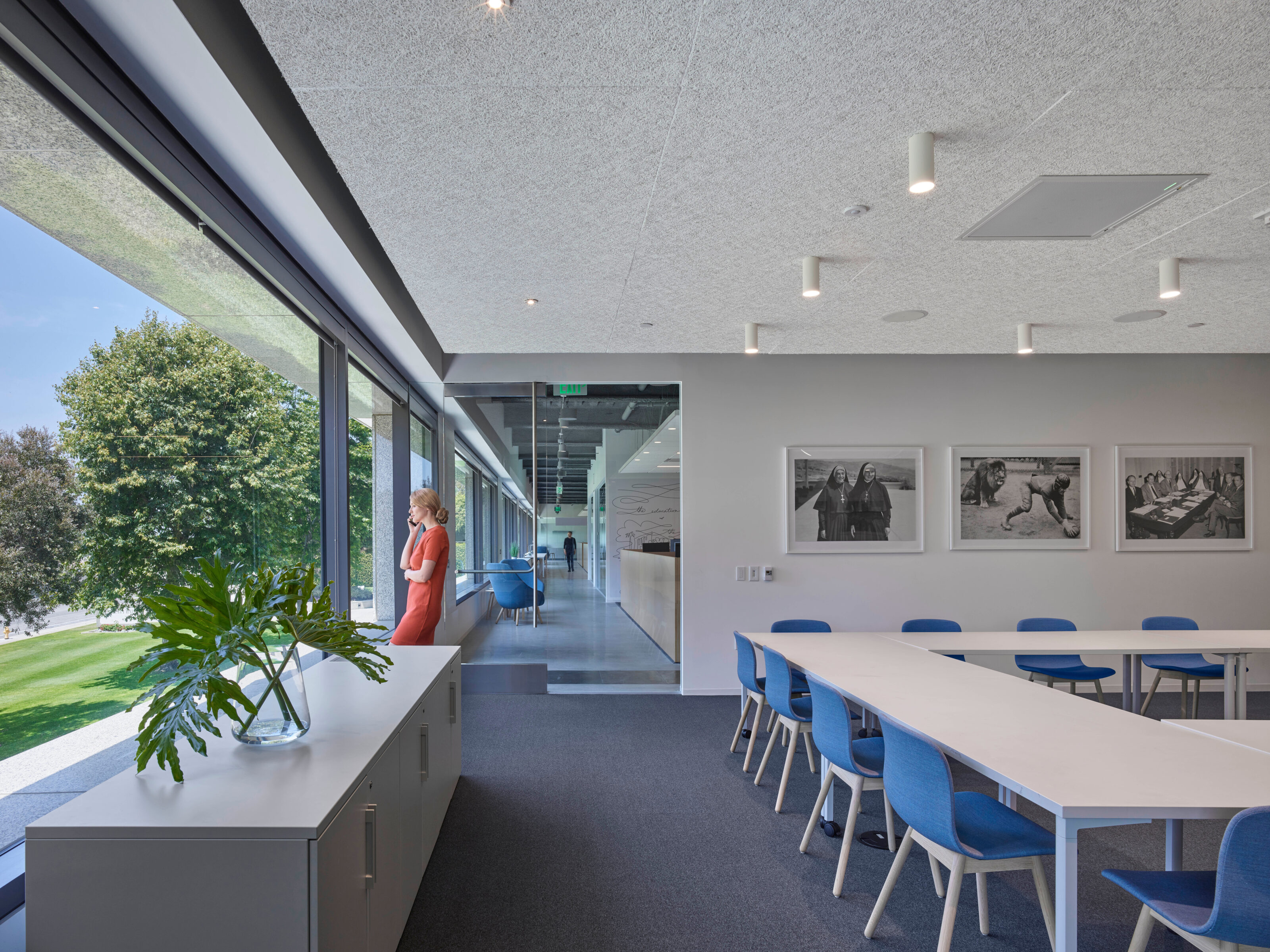 Blue chairs surround a white meeting table in the assembly room where a woman takes a call near the beautiful glass wall and exit.