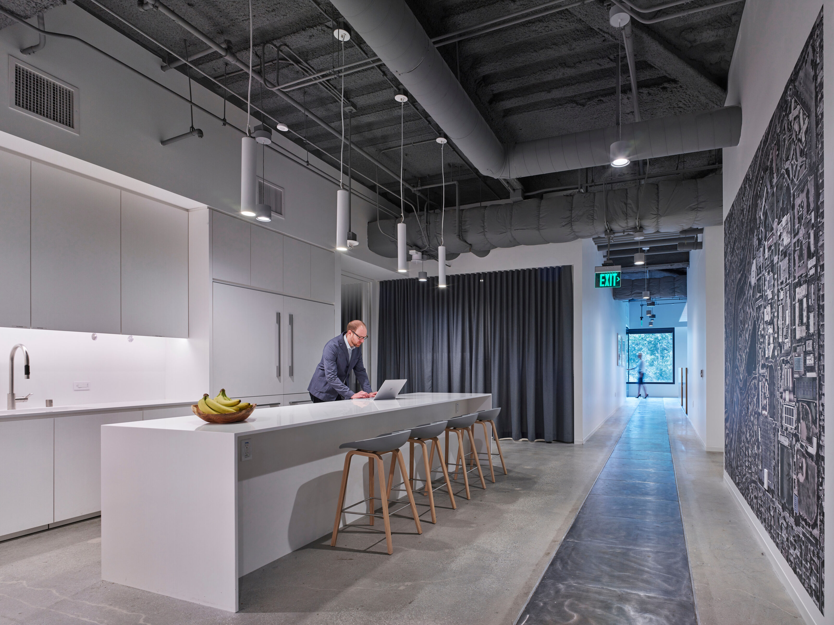 A modern white break room with a sleek white bar table and gray stools featuring wooden legs, where a man works on his laptop at the high table.