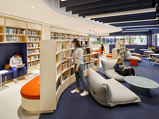 In the open reading area, custom bookshelf booths offer cozy seating along with lounge chairs and coffee tables, all situated on a dark blue carpet.