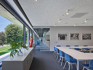 Blue chairs surround a white meeting table in the assembly room where a woman takes a call near the beautiful glass wall and exit.