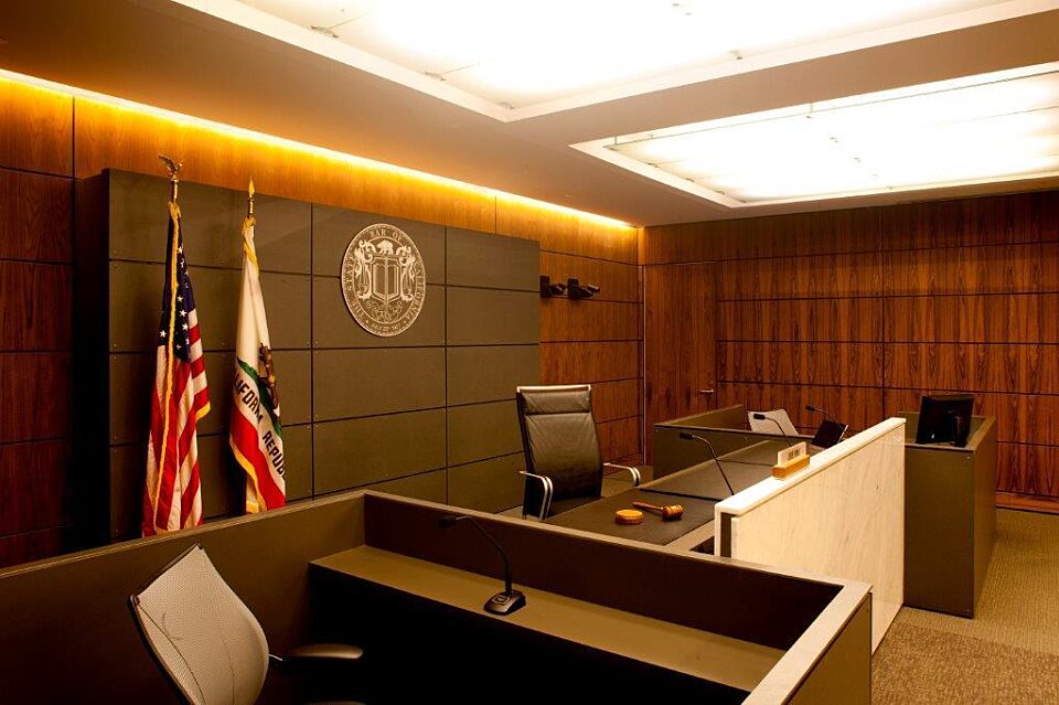 A courtroom with two desks and chairs, featuring a wooden floor, prominent lighting, and the presence of an American flag.