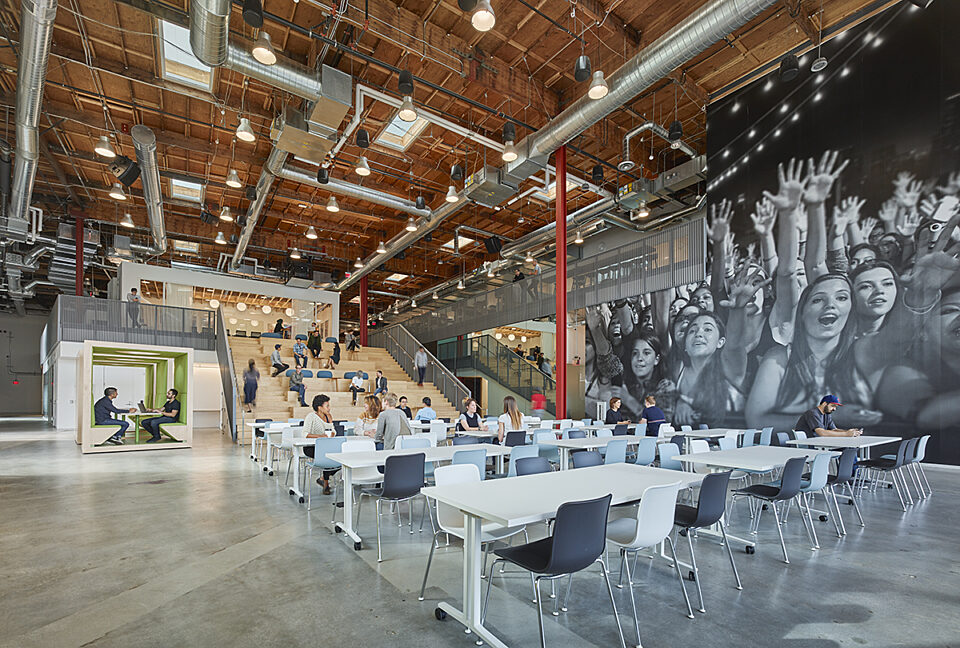 In an open area, there are white conference tables and white and black desk chairs. There is a wooden stair-style resting area in the back.